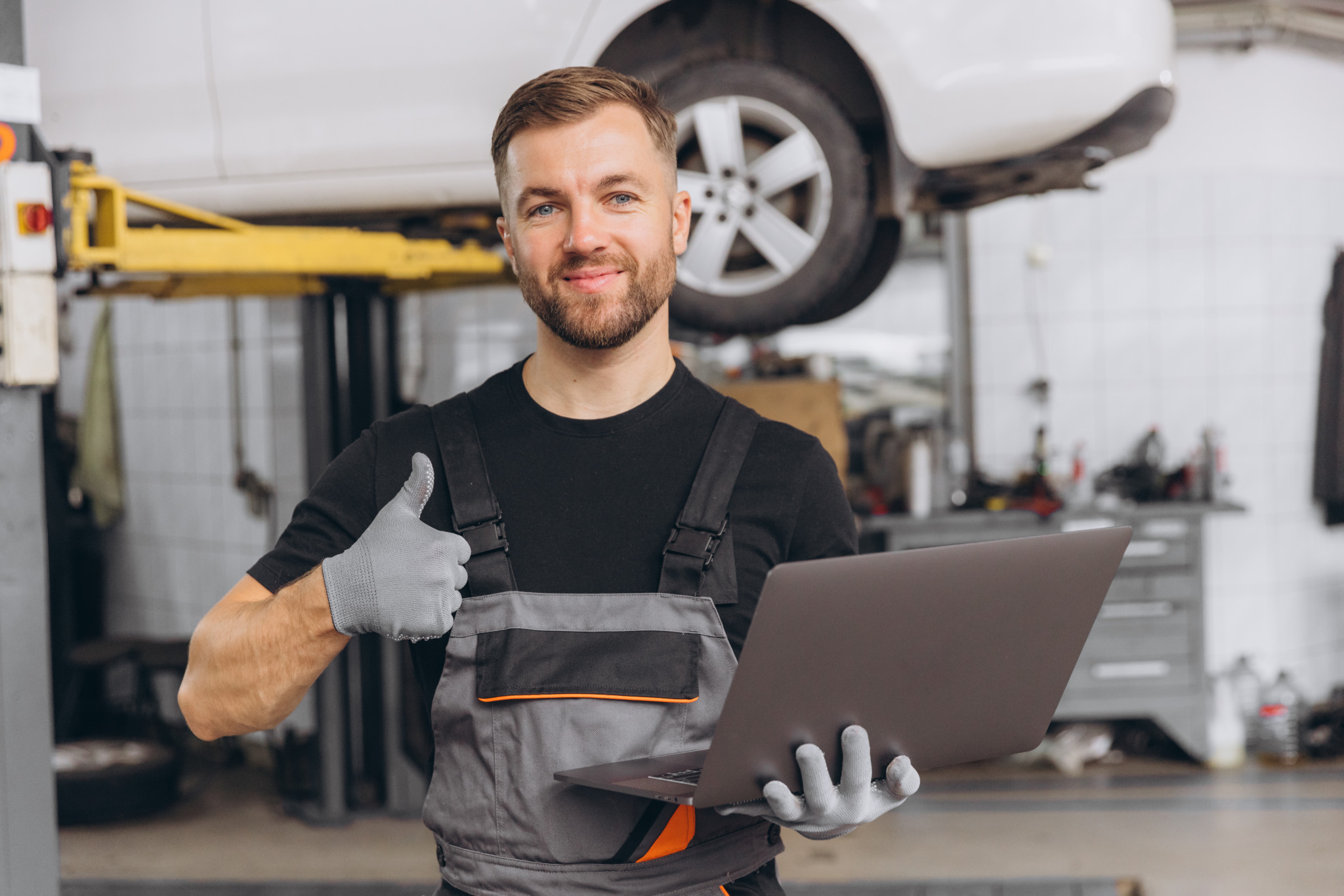 Worker of autoservice, smiling bearded man holding laptop in hands and showing thumbs up. Mechanic fixing, repairing automobile lifted behind.